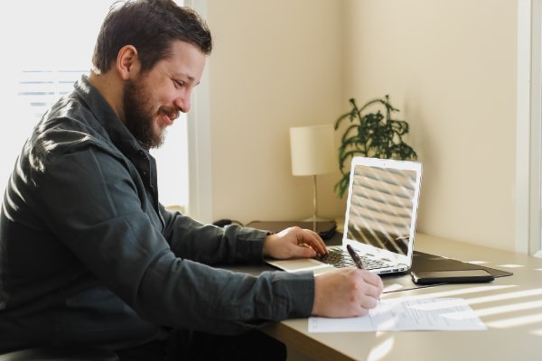 Caucasian smiling male manager filling documents and working with laptop. Concept of writing application and office worker job. Bearded man working with documents and computer.
