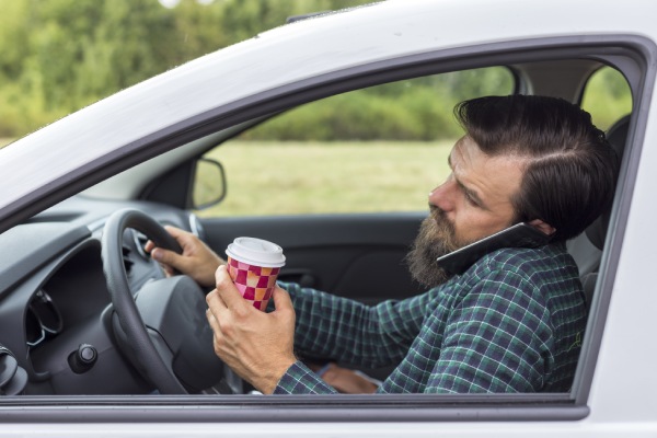 Young man speaking on phone and drinking coffee while driving his car