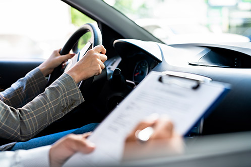 A woman behind the wheel of a car, holding a clipboard, likely engaged in the Impact Texas Adult Drivers Course.