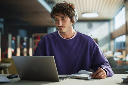  A man wearing headphones works at a table with a laptop, highlighting the convenience of Texas defensive driving online.