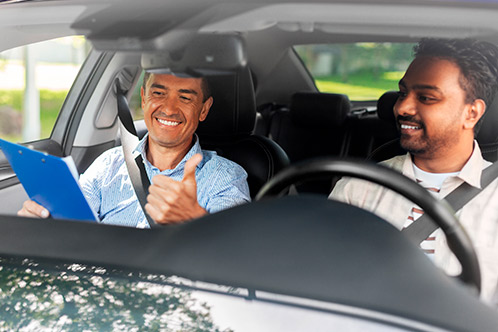 Two men in a car, one with a clipboard, both smiling and giving a thumbs up, encouraging Texas Adult Drivers Ed completion.
