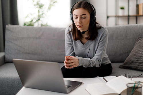A woman wearing headphones is seated on a couch, using her laptop to research Texas defensive driving insurance savings.