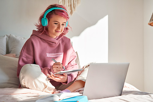 A woman wearing headphones sits on a bed, engaged with her laptop for a Spring Break project on driving permits.