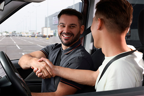 Inside a car, two men shake hands, highlighting the Spring Break Project for preparing teens for their driving permits.