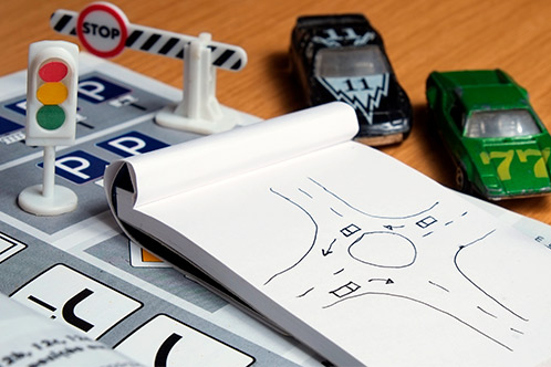 Toy cars and road signs surround a notepad with a roundabout sketch on a table. The setup resembles a traffic planning scene.