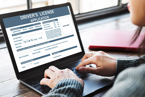 A person types on a laptop displaying a driver's license application form. They appear focused, with a red notebook nearby, suggesting a work setting.