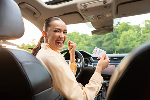 A woman in a car celebrates joyfully, holding a driver's license. She's smiling broadly, wearing a light blouse.