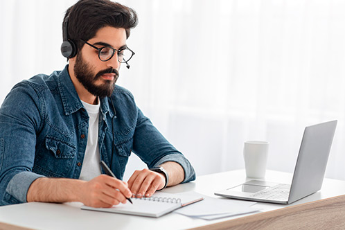 A bearded man wearing glasses works at a desk with a laptop, engaged in an online Texas driver education session.