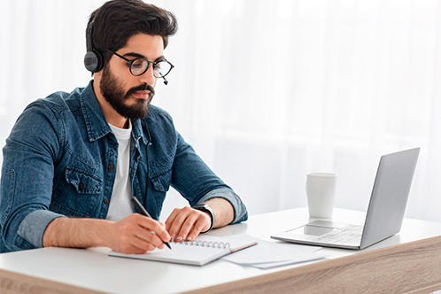 A bearded man wearing glasses is seated at a desk, engaged with his laptop.