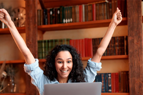 Yes, I'm A Winner. Excited emotional ecstatic lady celebrating success, victory or great news sitting at table, using personal computer, screaming yeah and raising hands up. Smiling happy woman in joy