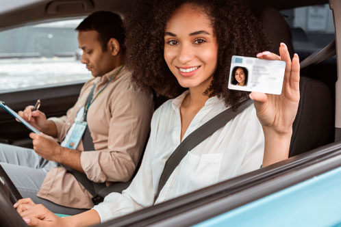 Joyful Middle Eastern Woman Showcasing Her Driving License To Camera Through Car Window Sitting Inside, While Professional Instructor Taking Notes After Successful Test Drive. Selective Focus