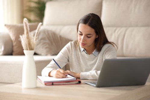 Student studying taking notes on notebook beside a laptop at home