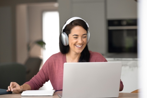 Smiling woman in headphones listening audio online course, engaged in professional, educational webinar event seated at table with laptop in cozy home interior. On-line studying, e-learning, tuition